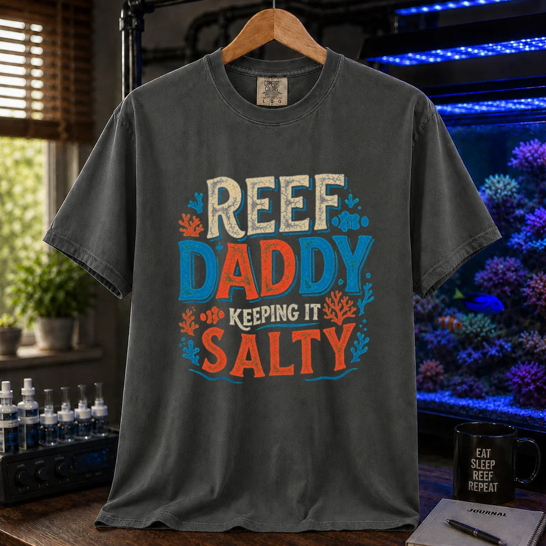 Saltwater aquarium graphic t-shirt design featuring bold distressed typography reading Reef Daddy Keeping It Salty with accent illustrations of clownfish and coral silhouettes on a dark grey tee