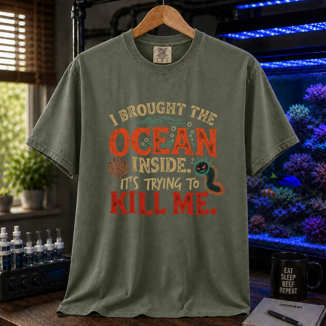 Saltwater aquarium graphic t-shirt design featuring distressed typography reading I Brought The Ocean Inside. It's Trying To Kill Me with a menacing bristle worm and coral illustration on a military green tee