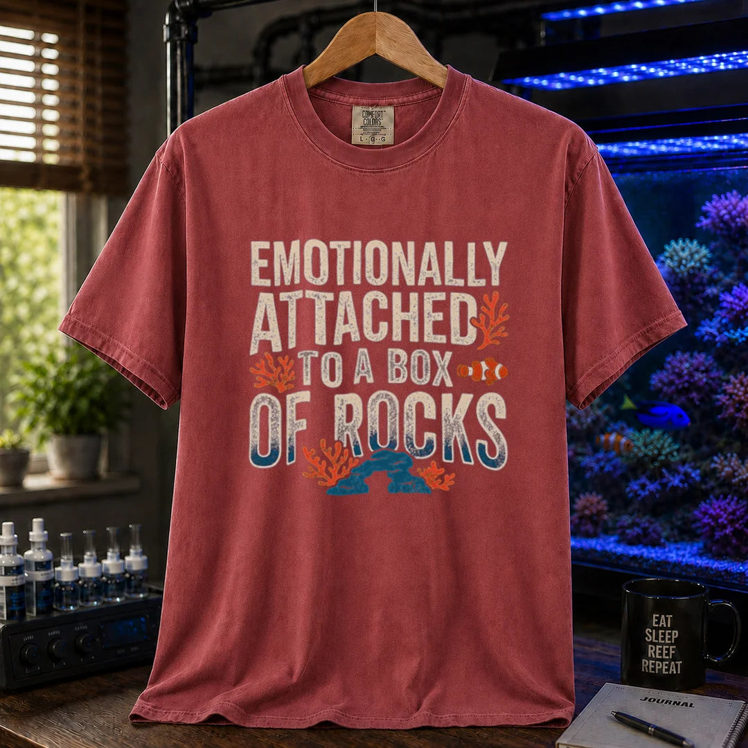 Saltwater aquarium graphic t-shirt featuring distressed block typography reading Emotionally Attached To A Box Of Rocks with coral and clownfish illustrations printed on a maroon, red tee displayed in a reef room setting