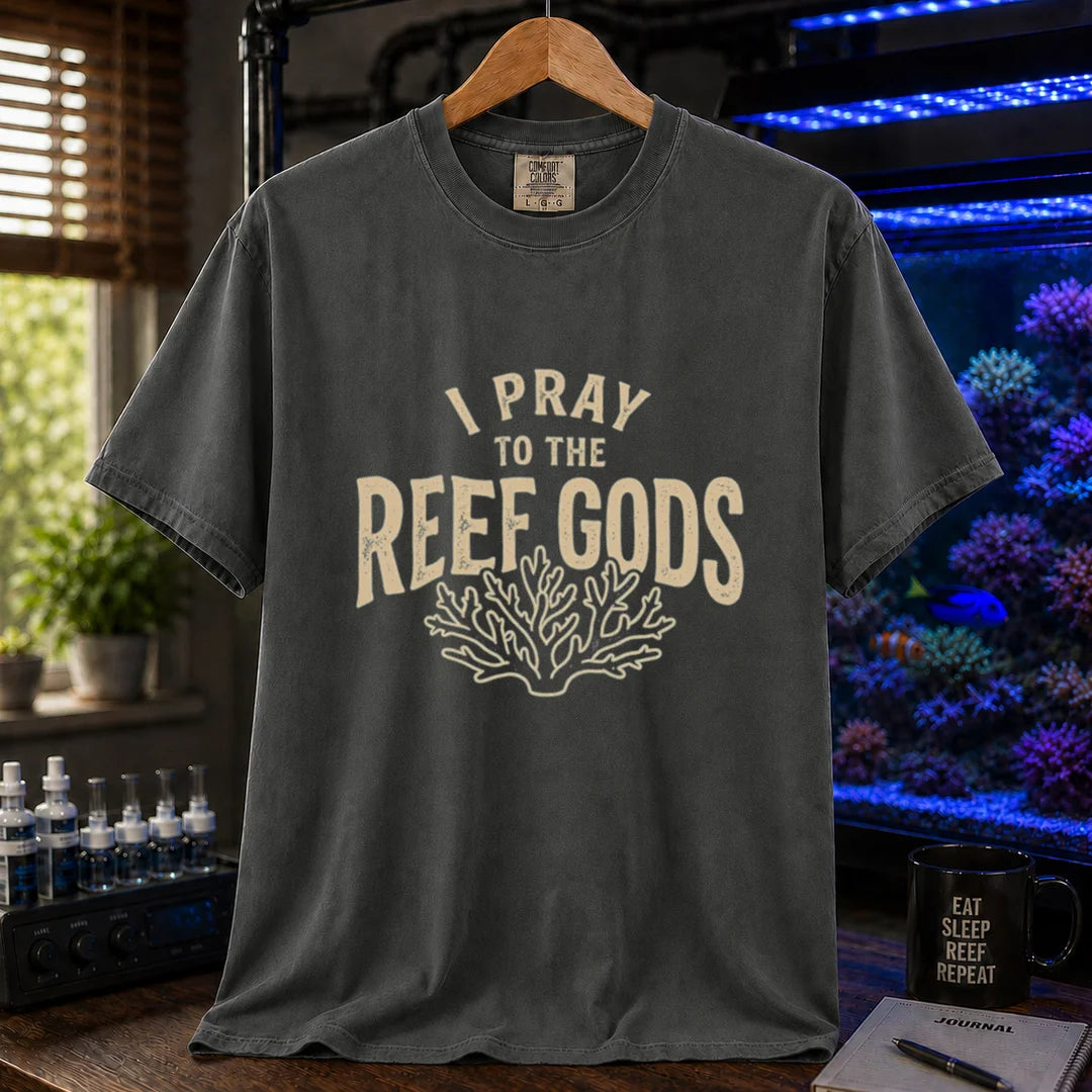 Saltwater aquarium graphic t-shirt featuring distressed cream typography reading I Pray To The Reef Gods with a coral line art graphic printed on a dark grey tee displayed in a reef room setting