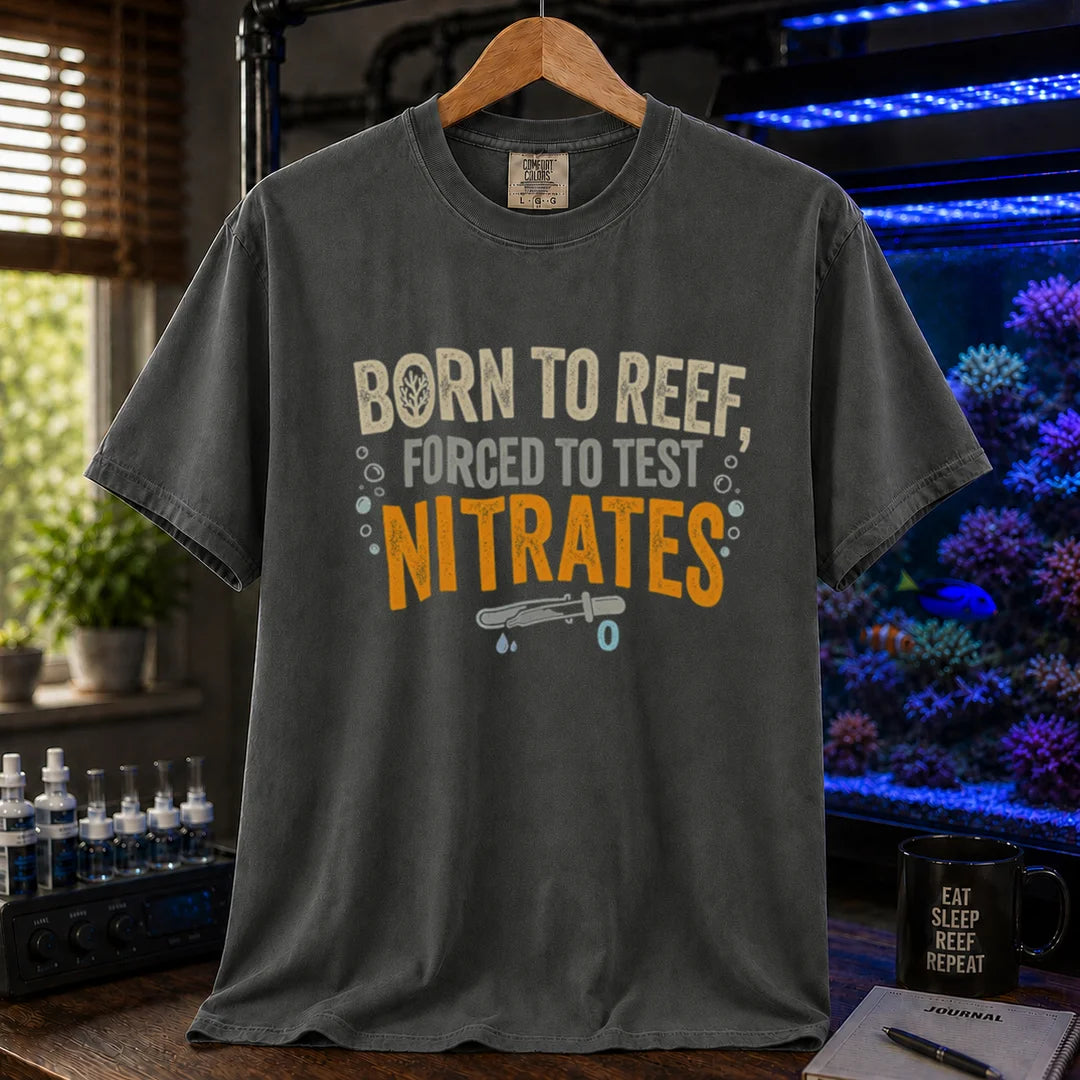 Saltwater aquarium graphic t-shirt featuring distressed cream, gray, and yellow-orange typography reading Born To Reef, Forced To Test Nitrates with a test vial icon printed on a dark grey tee displayed in a reef room setting