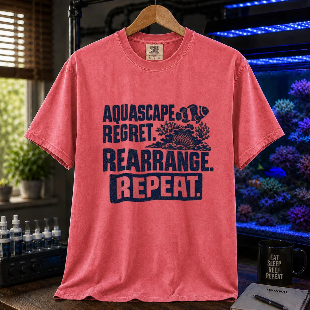 Saltwater aquarium graphic t-shirt featuring distressed dark blue typography reading Aquascape. Regret. Rearrange. Repeat. with a coral and clownfish illustration printed on a watermelon tee displayed in a reef room setting
