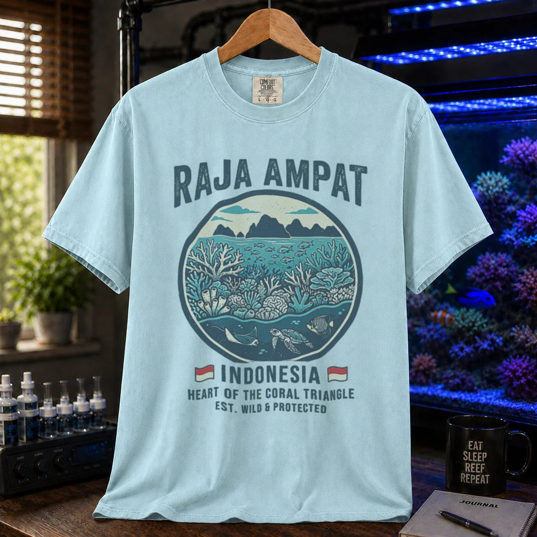 Saltwater aquarium graphic t-shirt featuring a circular reef landscape illustration and text: RAJA AMPAT INDONESIA HEART OF THE CORAL TRIANGLE EST. WILD & PROTECTED, light blue crew neck tee