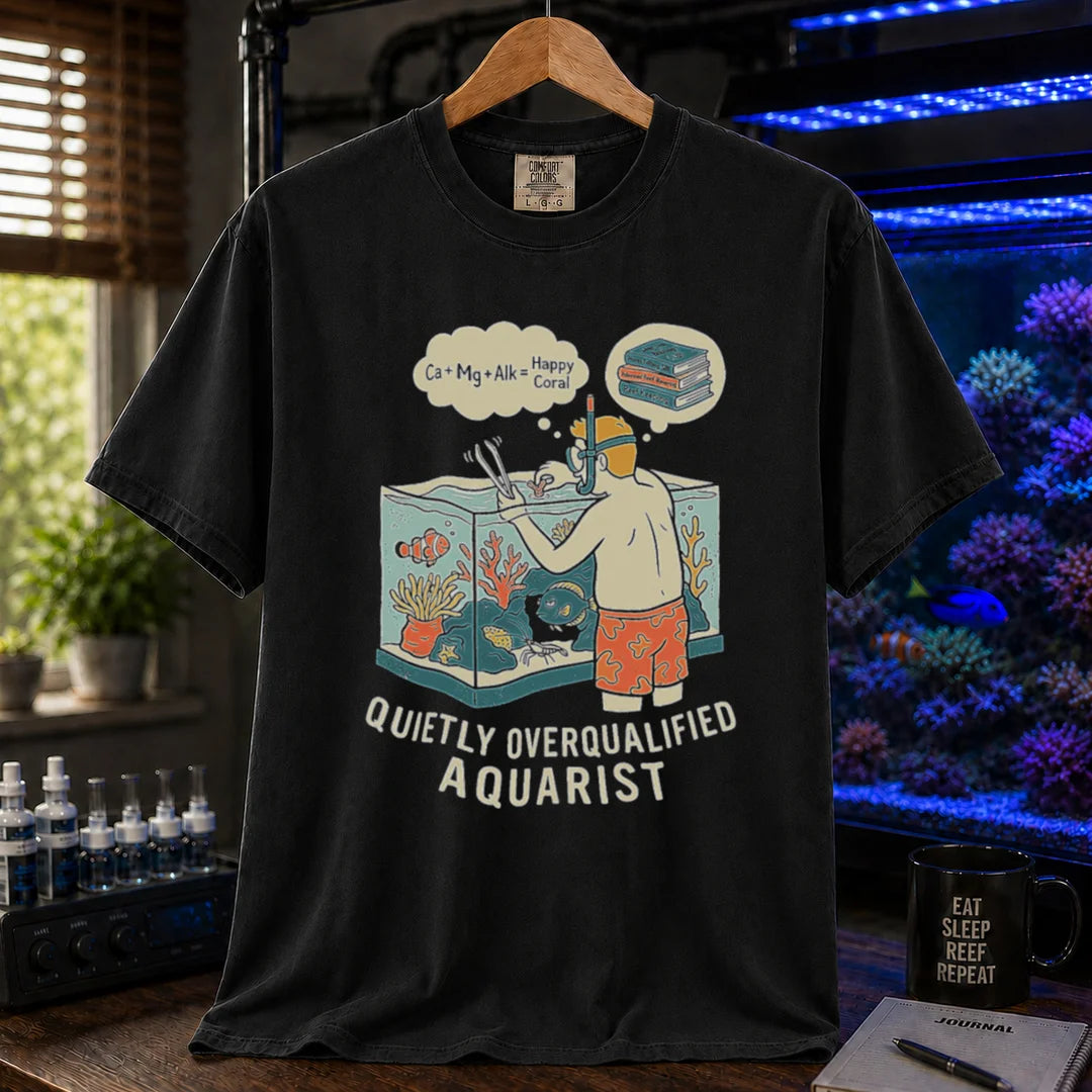 Saltwater aquarium graphic t-shirt featuring an aquarist maintaining a tank with chemical equation thought bubbles and text: QUIETLY OVERQUALIFIED AQUARIST, black crew neck tee