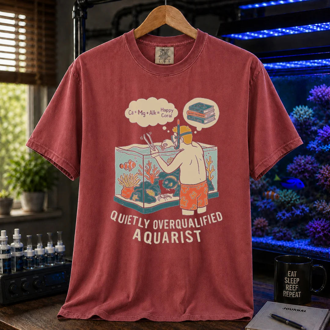 Saltwater aquarium graphic t-shirt featuring an aquarist maintaining a tank with chemical equation thought bubbles and text: QUIETLY OVERQUALIFIED AQUARIST, maroon, red crew neck tee