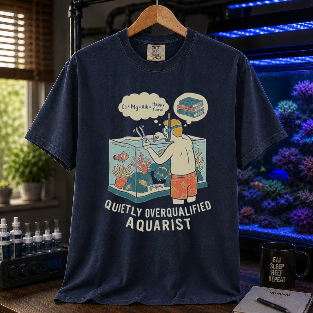Saltwater aquarium graphic t-shirt featuring an aquarist maintaining a tank with chemical equation thought bubbles and text: QUIETLY OVERQUALIFIED AQUARIST, navy crew neck tee