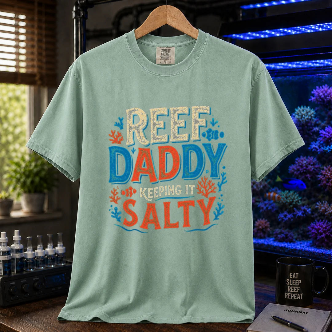 Saltwater aquarium graphic t-shirt design featuring bold distressed typography reading Reef Daddy Keeping It Salty with accent illustrations of clownfish and coral silhouettes on a light green tee