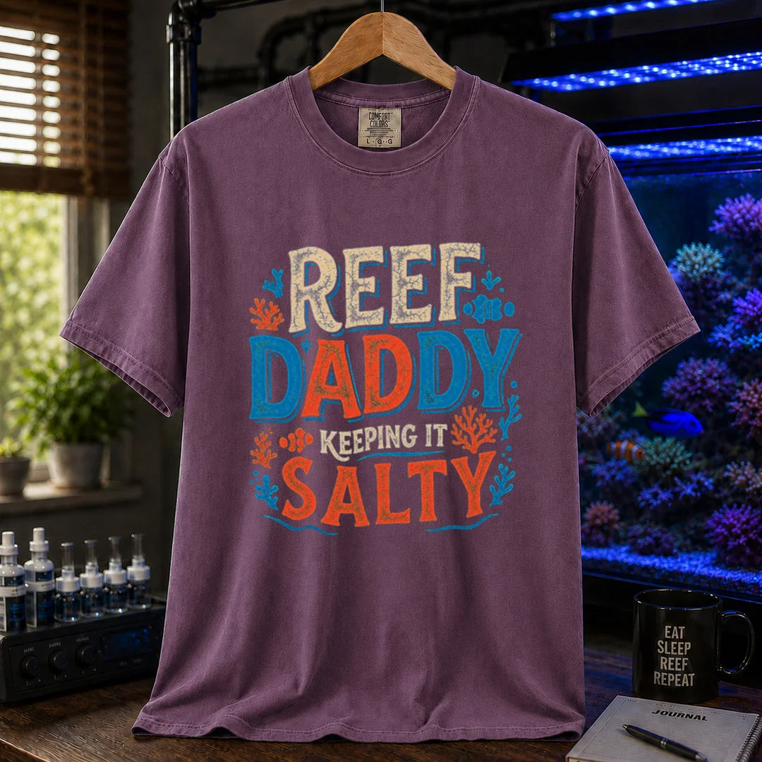 Saltwater aquarium graphic t-shirt design featuring bold distressed typography reading Reef Daddy Keeping It Salty with accent illustrations of clownfish and coral silhouettes on a purple tee