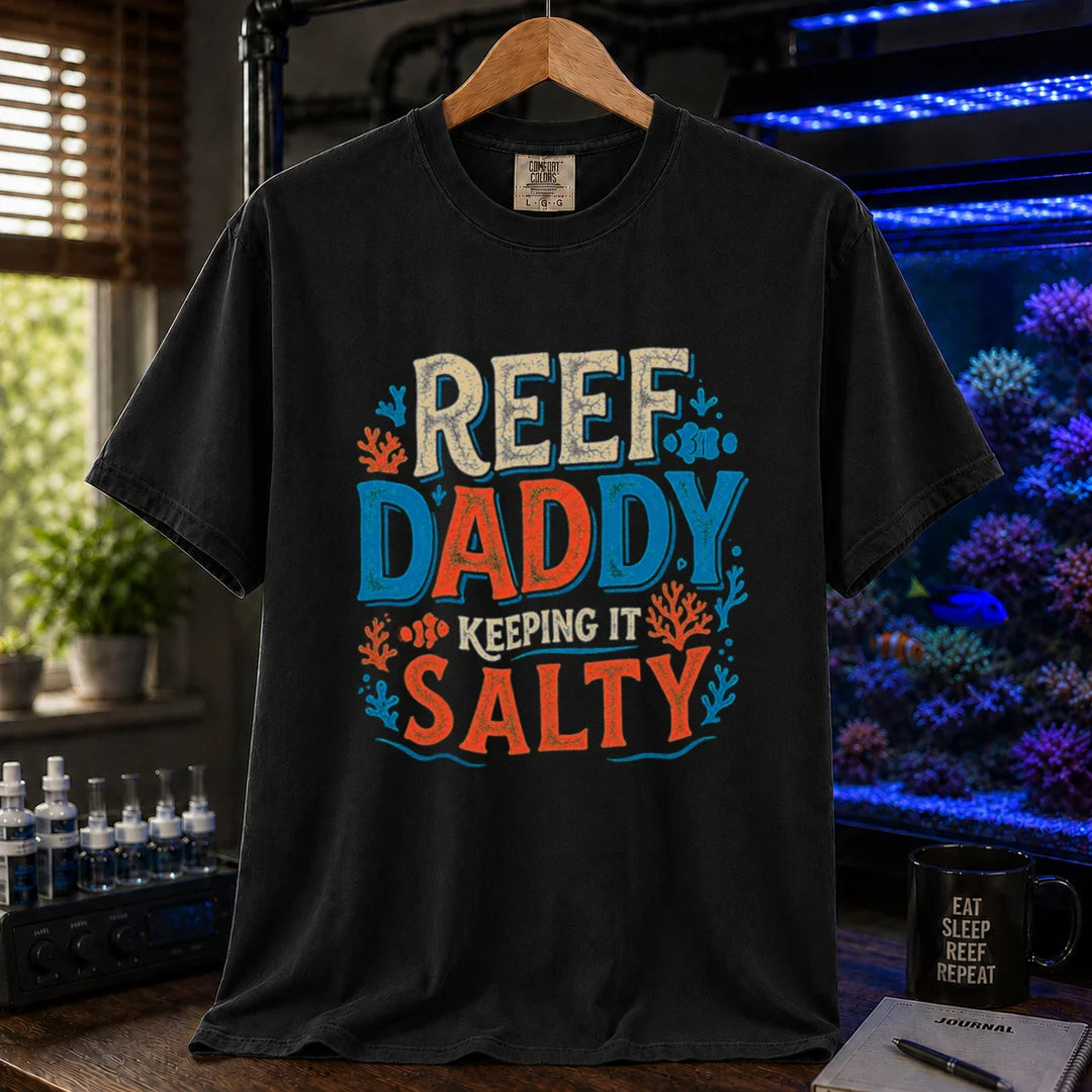 Saltwater aquarium graphic t-shirt design featuring bold distressed typography reading Reef Daddy Keeping It Salty with accent illustrations of clownfish and coral silhouettes on a BLACK tee