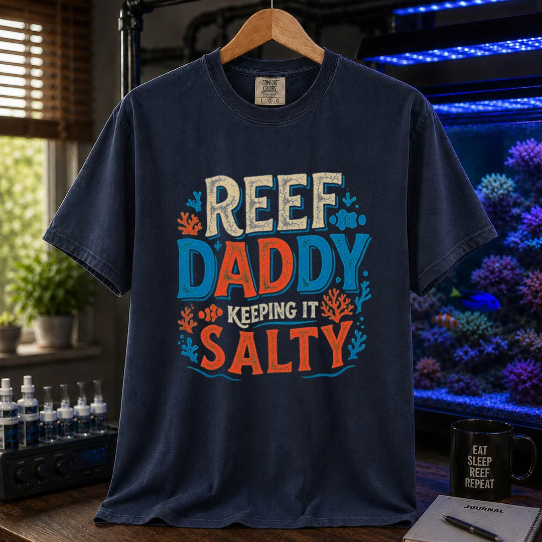 Saltwater aquarium graphic t-shirt design featuring bold distressed typography reading Reef Daddy Keeping It Salty with accent illustrations of clownfish and coral silhouettes on a navy tee
