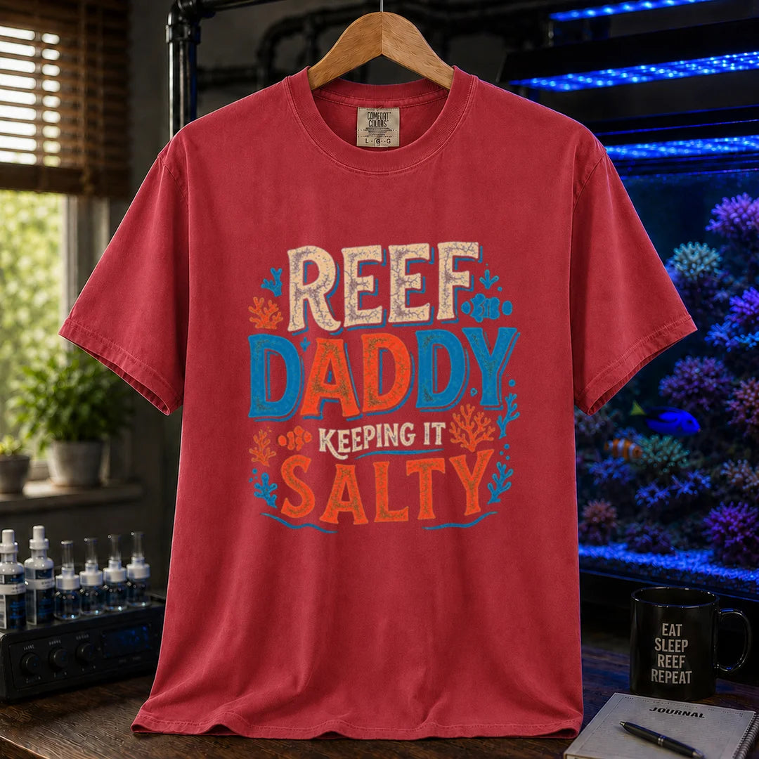 Saltwater aquarium graphic t-shirt design featuring bold distressed typography reading Reef Daddy Keeping It Salty with accent illustrations of clownfish and coral silhouettes on a red tee
