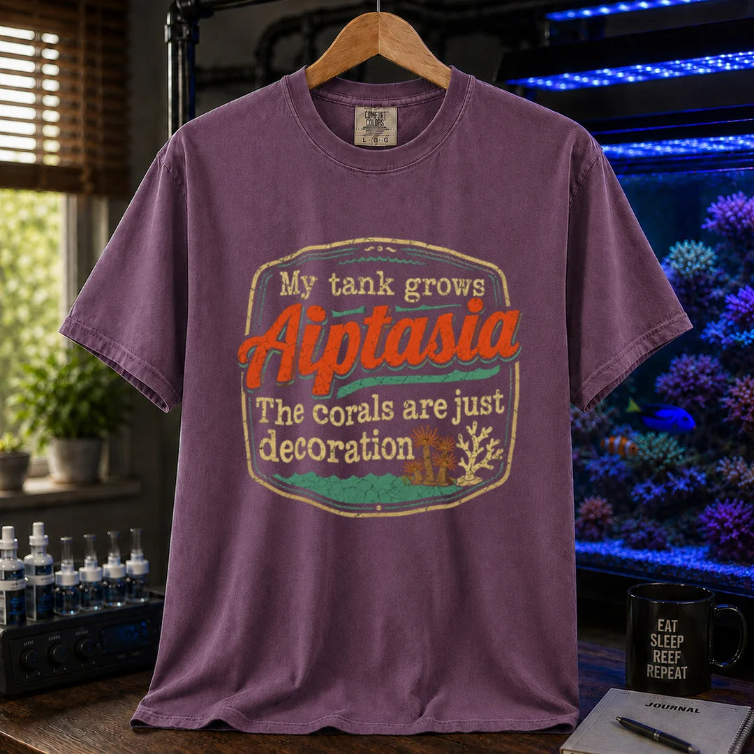 Saltwater aquarium graphic t-shirt design featuring a vintage badge reading My Tank Grows Aiptasia. The corals are just decoration with pest anemone illustrations on a purple tee