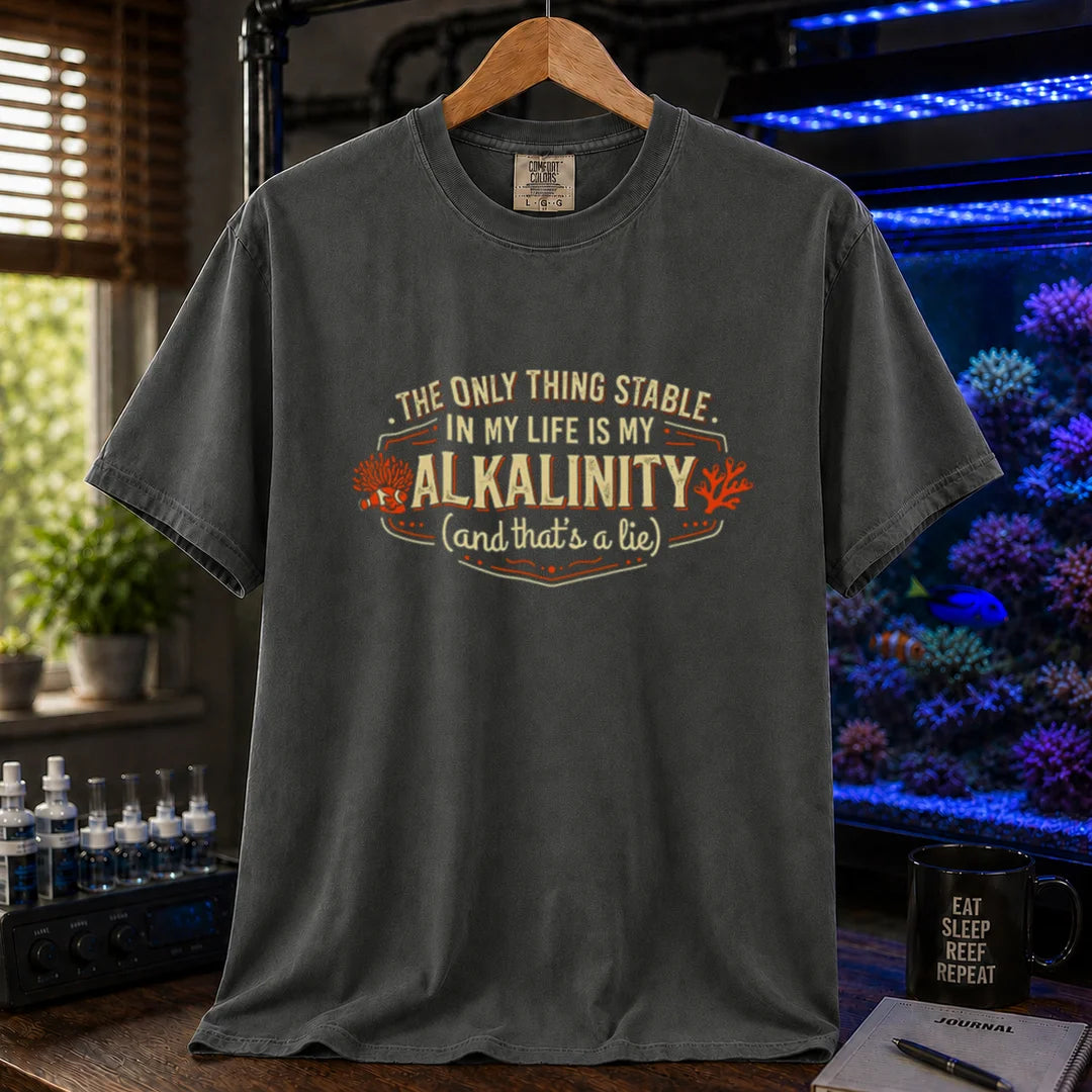 Saltwater aquarium graphic t-shirt design featuring a vintage badge reading The Only Thing Stable In My Life Is My Alkalinity (and that's a lie) with coral accents on a dark grey tee