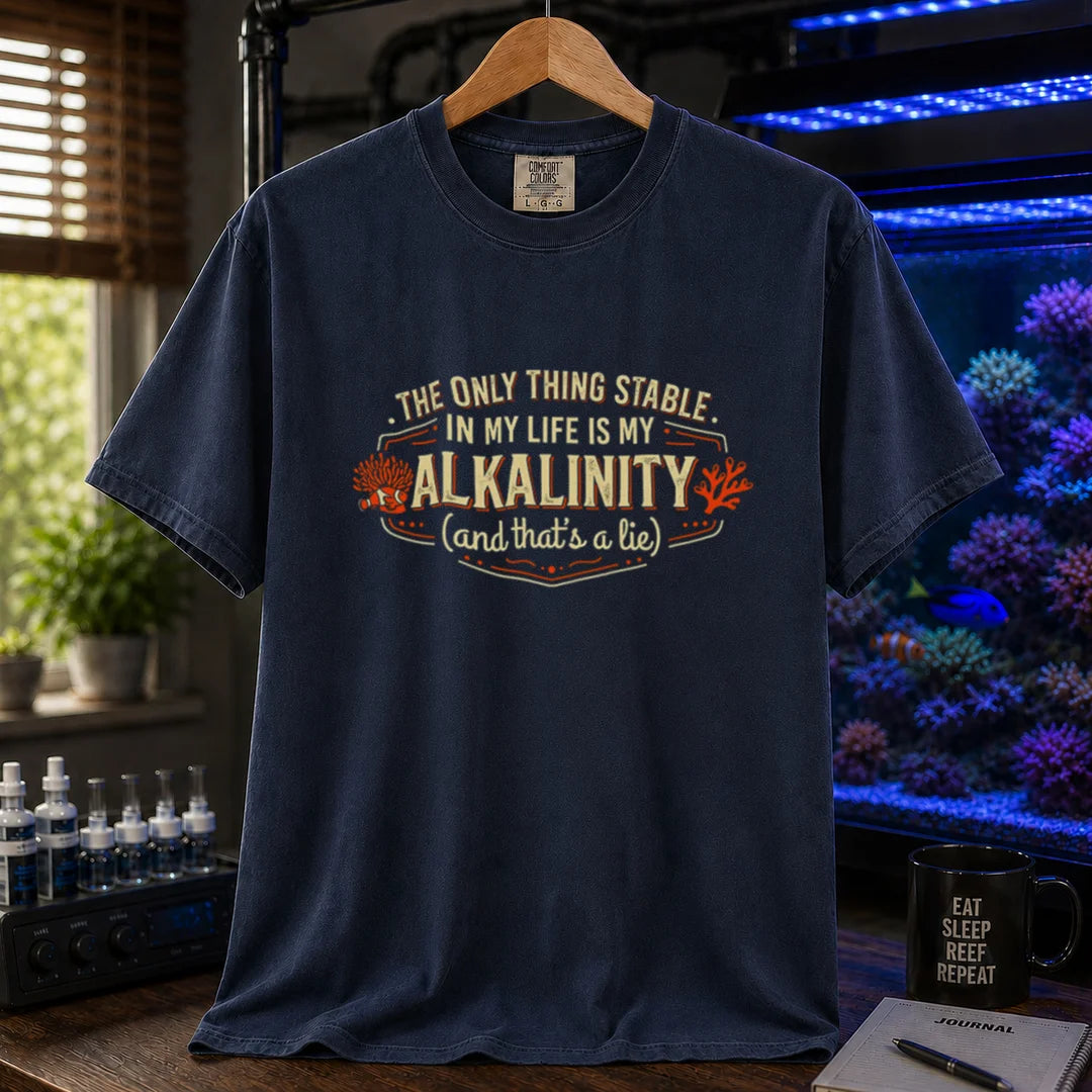 Saltwater aquarium graphic t-shirt design featuring a vintage badge reading The Only Thing Stable In My Life Is My Alkalinity (and that's a lie) with coral accents on a NAVY tee