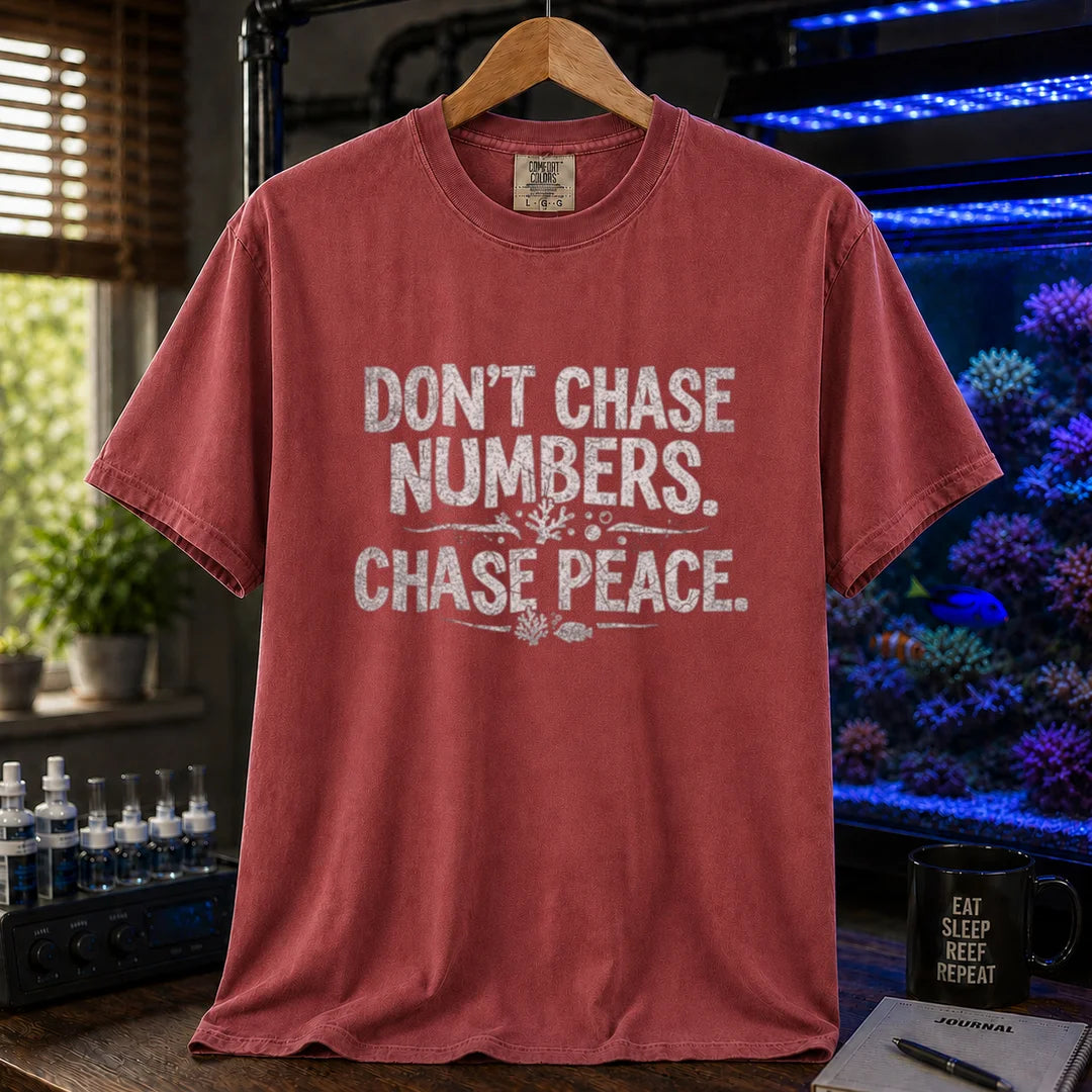 Saltwater aquarium graphic t-shirt design featuring distressed white typography reading Don't Chase Numbers. Chase Peace. with small coral and fish accents on a maroon, red tee
