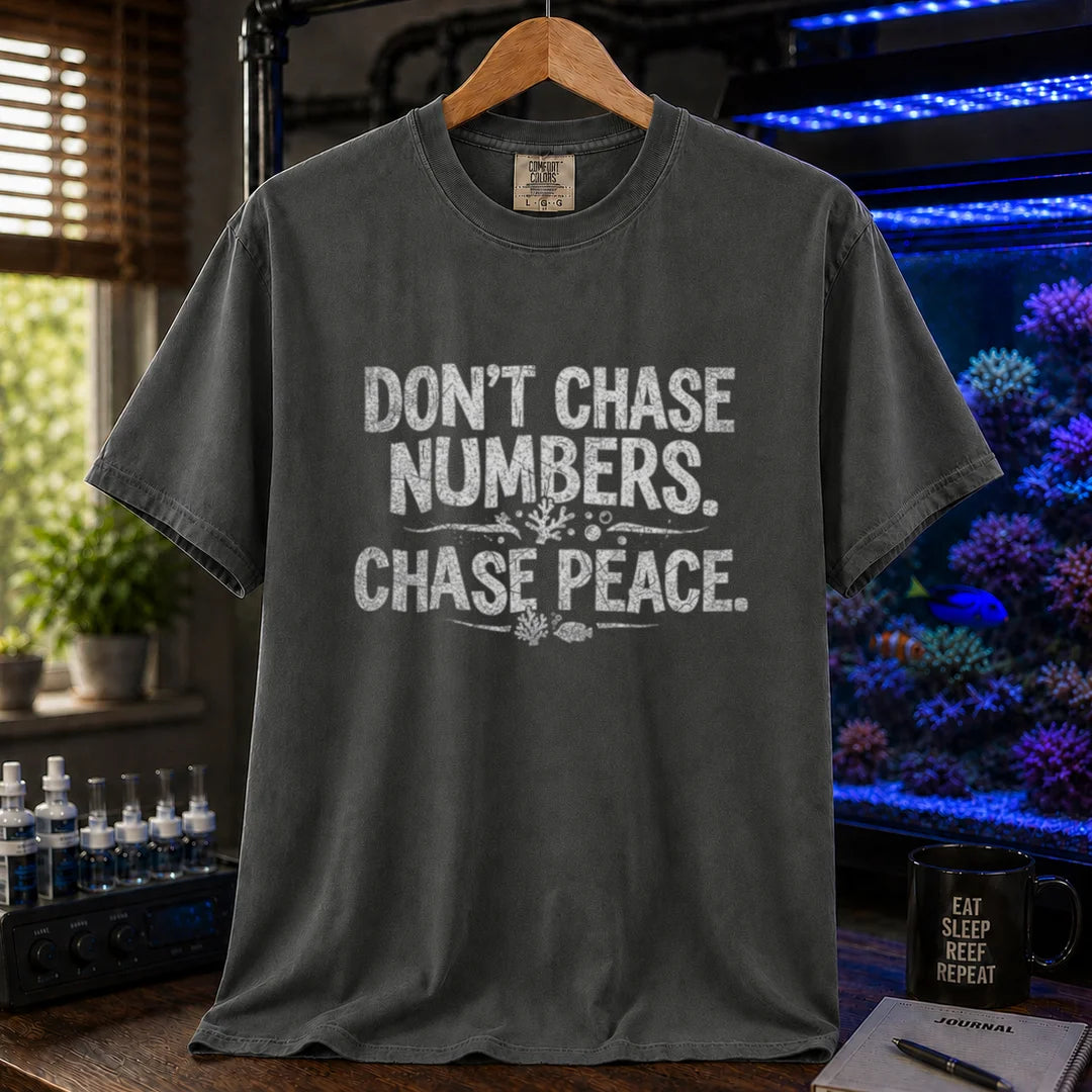 Saltwater aquarium graphic t-shirt design featuring distressed white typography reading Don't Chase Numbers. Chase Peace. with small coral and fish accents on a dark grey tee