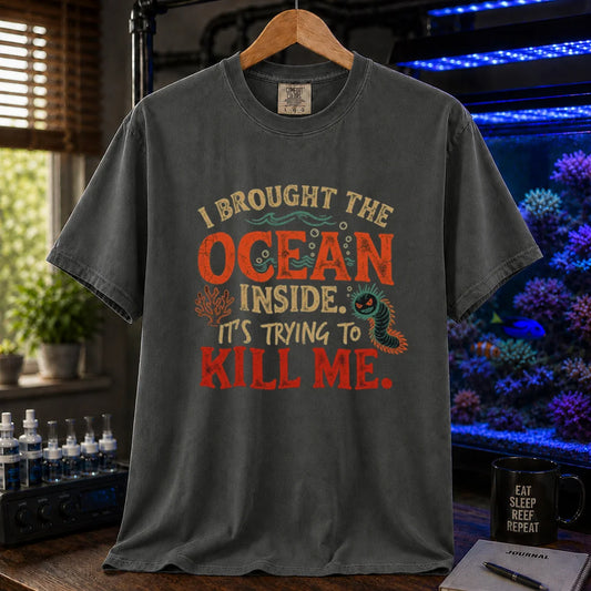 Saltwater aquarium graphic t-shirt design featuring distressed typography reading I Brought The Ocean Inside. It's Trying To Kill Me with a menacing bristle worm and coral illustration on a dark grey tee
