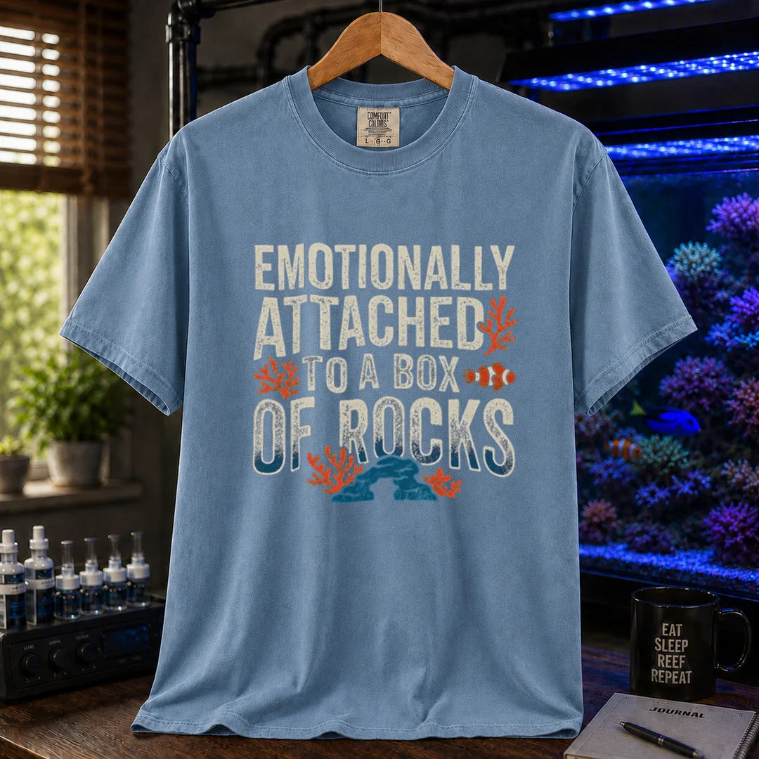 Saltwater aquarium graphic t-shirt featuring distressed block typography reading Emotionally Attached To A Box Of Rocks with coral and clownfish illustrations printed on a blue tee displayed in a reef room setting