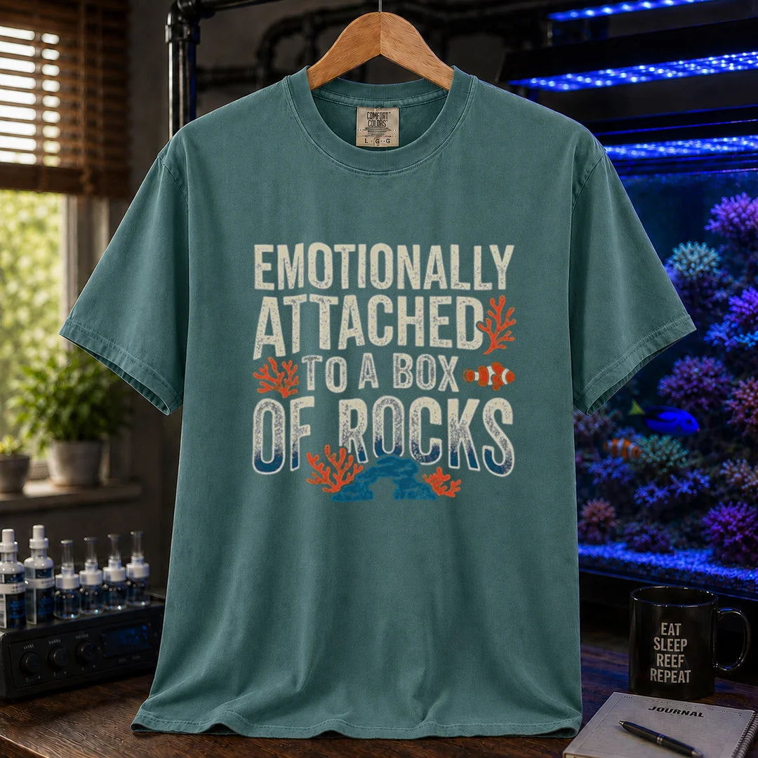 Saltwater aquarium graphic t-shirt featuring distressed block typography reading Emotionally Attached To A Box Of Rocks with coral and clownfish illustrations printed on a green tee displayed in a reef room setting