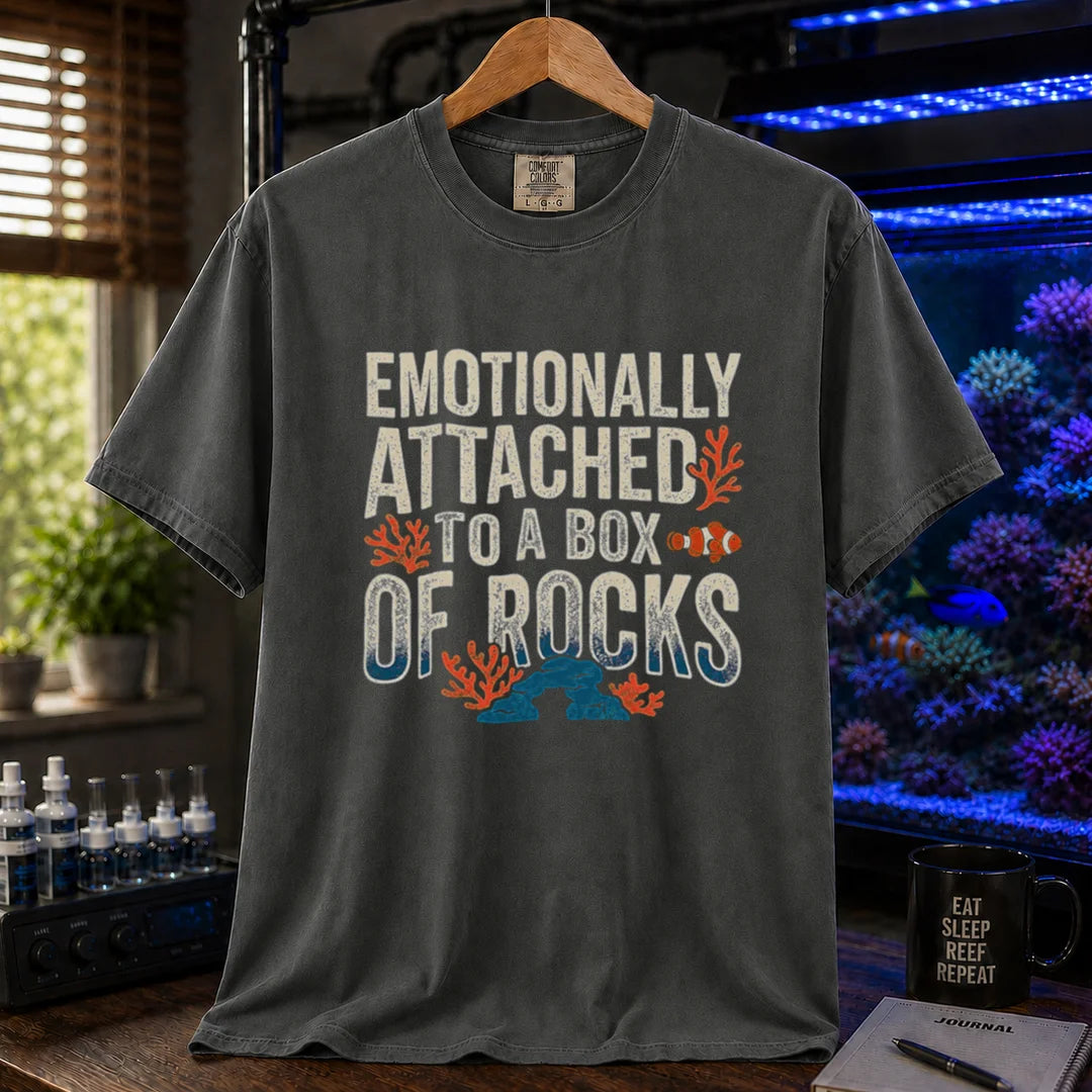 Saltwater aquarium graphic t-shirt featuring distressed block typography reading Emotionally Attached To A Box Of Rocks with coral and clownfish illustrations printed on a dark grey tee displayed in a reef room setting