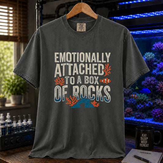 Saltwater aquarium graphic t-shirt featuring distressed block typography reading Emotionally Attached To A Box Of Rocks with coral and clownfish illustrations printed on a dark grey tee displayed in a reef room setting
