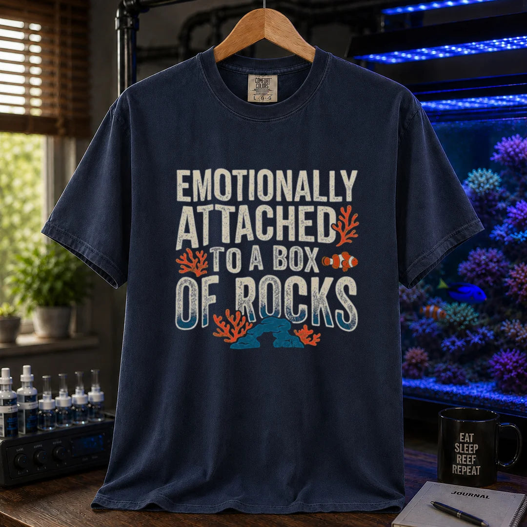 Saltwater aquarium graphic t-shirt featuring distressed block typography reading Emotionally Attached To A Box Of Rocks with coral and clownfish illustrations printed on a navy tee displayed in a reef room setting