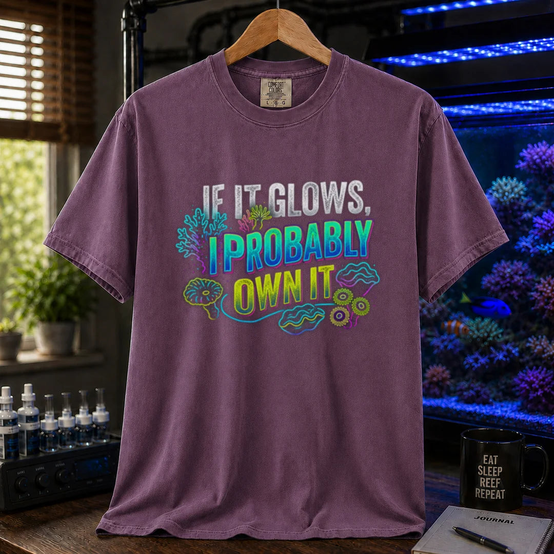 Saltwater aquarium graphic t-shirt featuring vibrant neon typography reading If It Glows, I Probably Own It with glowing fluorescent coral and clam graphics printed on a purple tee displayed in a reef room setting