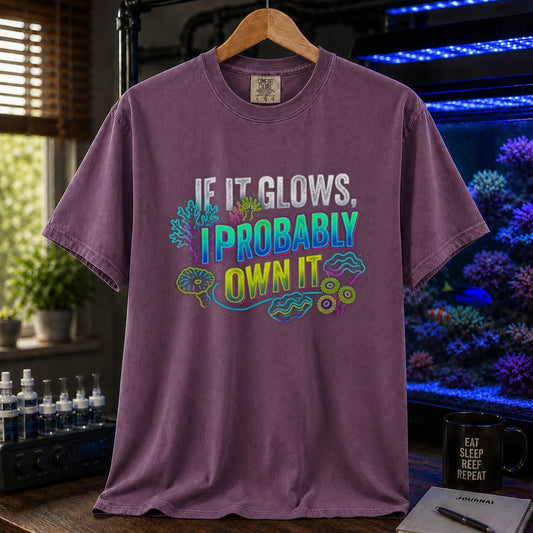 Saltwater aquarium graphic t-shirt featuring vibrant neon typography reading If It Glows, I Probably Own It with glowing fluorescent coral and clam graphics printed on a purple tee displayed in a reef room setting