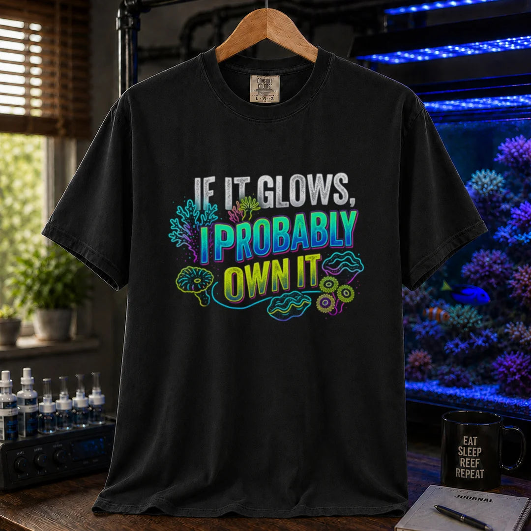 Saltwater aquarium graphic t-shirt featuring vibrant neon typography reading If It Glows, I Probably Own It with glowing fluorescent coral and clam graphics printed on a black tee displayed in a reef room setting