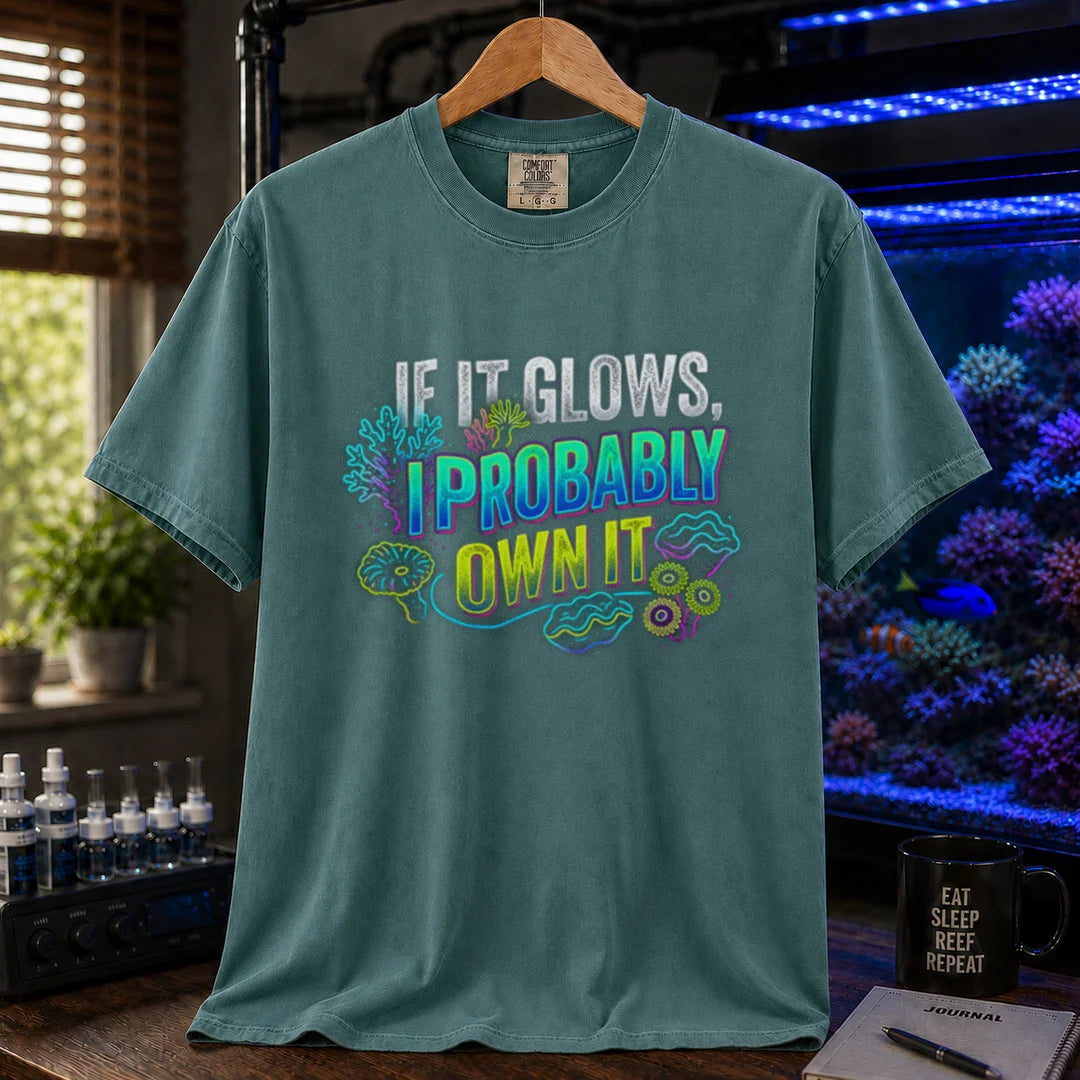Saltwater aquarium graphic t-shirt featuring vibrant neon typography reading If It Glows, I Probably Own It with glowing fluorescent coral and clam graphics printed on a green tee displayed in a reef room setting