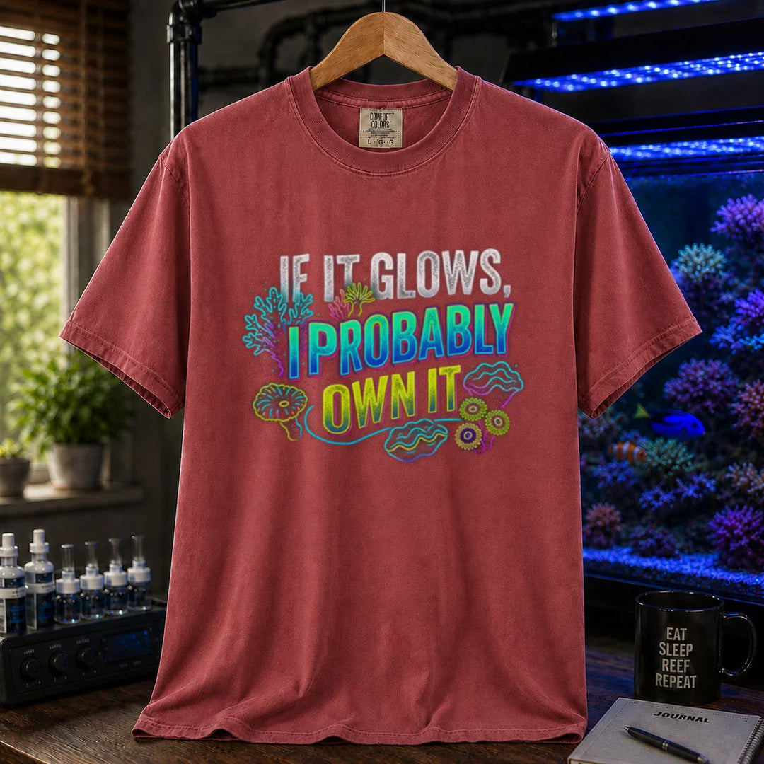 Saltwater aquarium graphic t-shirt featuring vibrant neon typography reading If It Glows, I Probably Own It with glowing fluorescent coral and clam graphics printed on a maroon, red tee displayed in a reef room setting