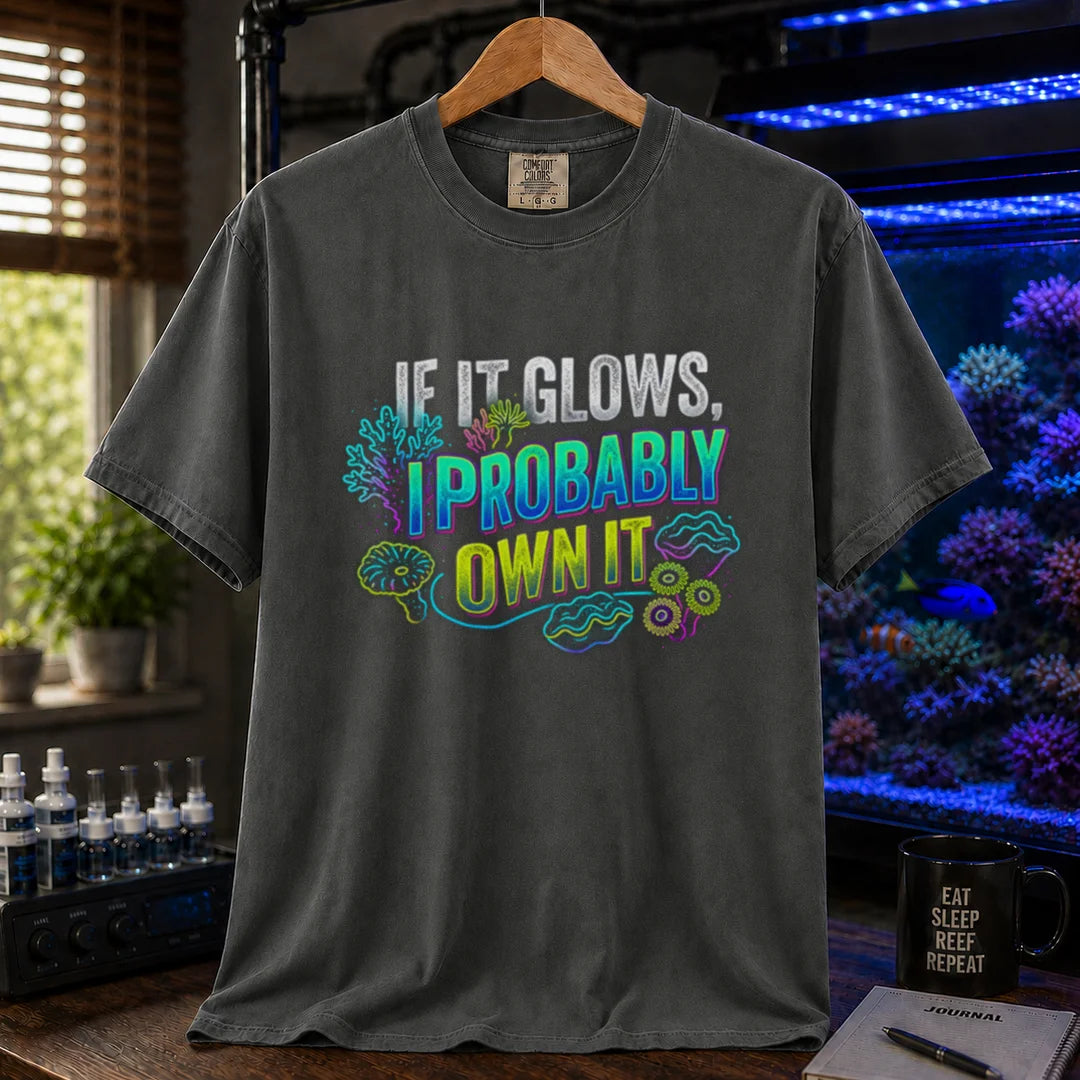 Saltwater aquarium graphic t-shirt featuring vibrant neon typography reading If It Glows, I Probably Own It with glowing fluorescent coral and clam graphics printed on a dark grey tee displayed in a reef room setting