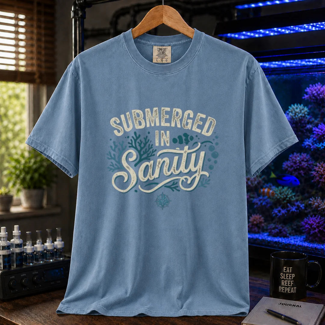 Saltwater aquarium graphic t-shirt featuring distressed cream and teal typography reading Submerged In Sanity with coral and bubble accents printed on a blue tee displayed in a reef room setting
