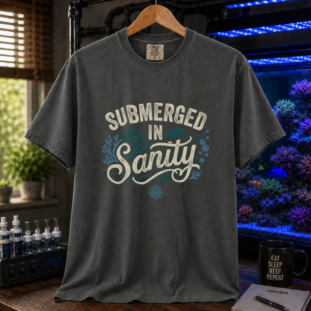 Saltwater aquarium graphic t-shirt featuring distressed cream and teal typography reading Submerged In Sanity with coral and bubble accents printed on a dark grey tee displayed in a reef room setting