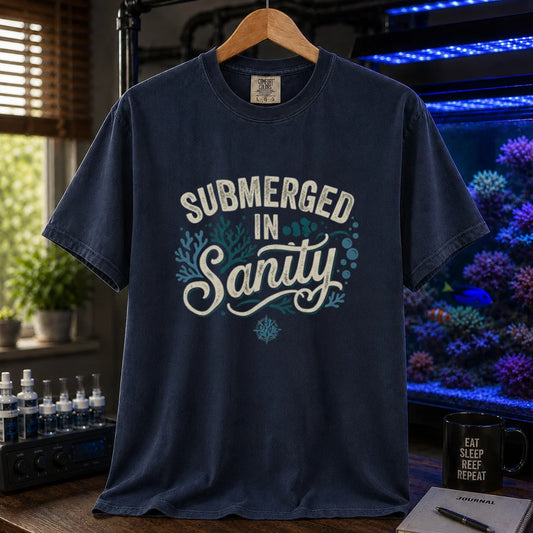 Saltwater aquarium graphic t-shirt featuring distressed cream and teal typography reading Submerged In Sanity with coral and bubble accents printed on a navy tee displayed in a reef room setting