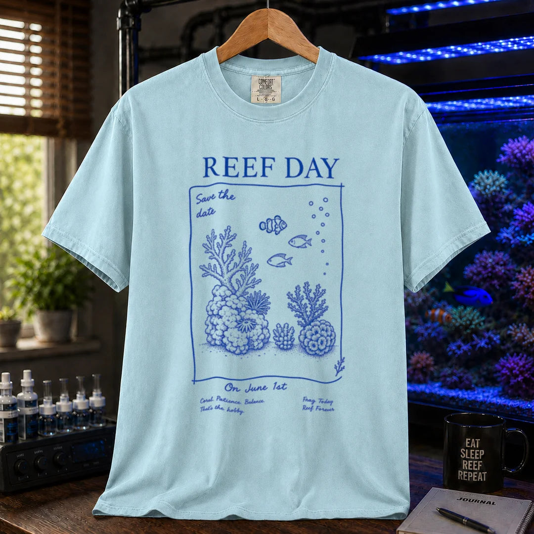 Saltwater aquarium graphic t-shirt featuring blue line-art typography and illustrations reading Reef Day Save The Date On June 1st with coral and fish graphics printed on a light blue tee displayed in a reef room setting