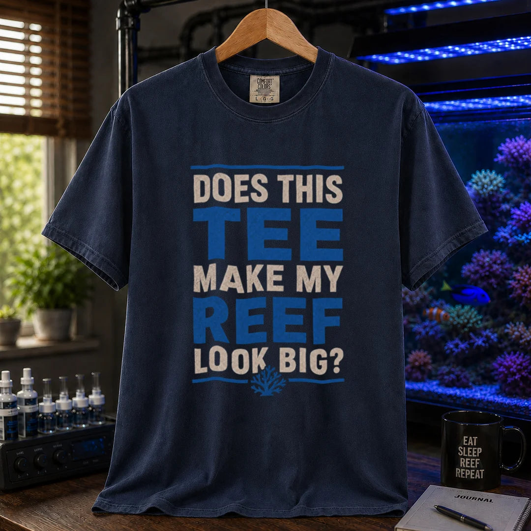 Saltwater aquarium graphic t-shirt featuring distressed white and blue block typography reading Does This Tee Make My Reef Look Big? with a small coral icon printed on a navy tee displayed in a reef room setting