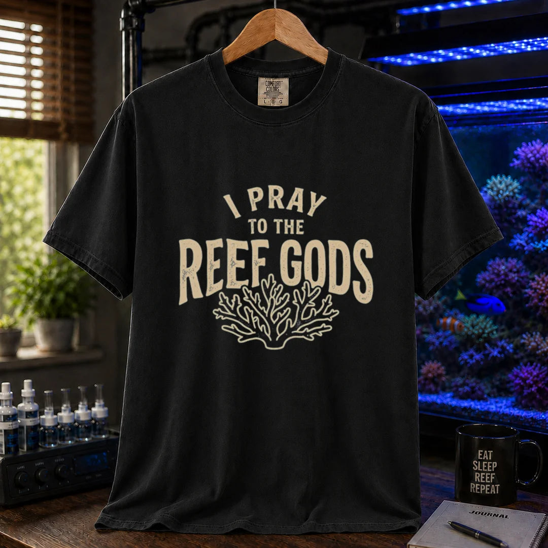 Saltwater aquarium graphic t-shirt featuring distressed cream typography reading I Pray To The Reef Gods with a coral line art graphic printed on a black tee displayed in a reef room setting