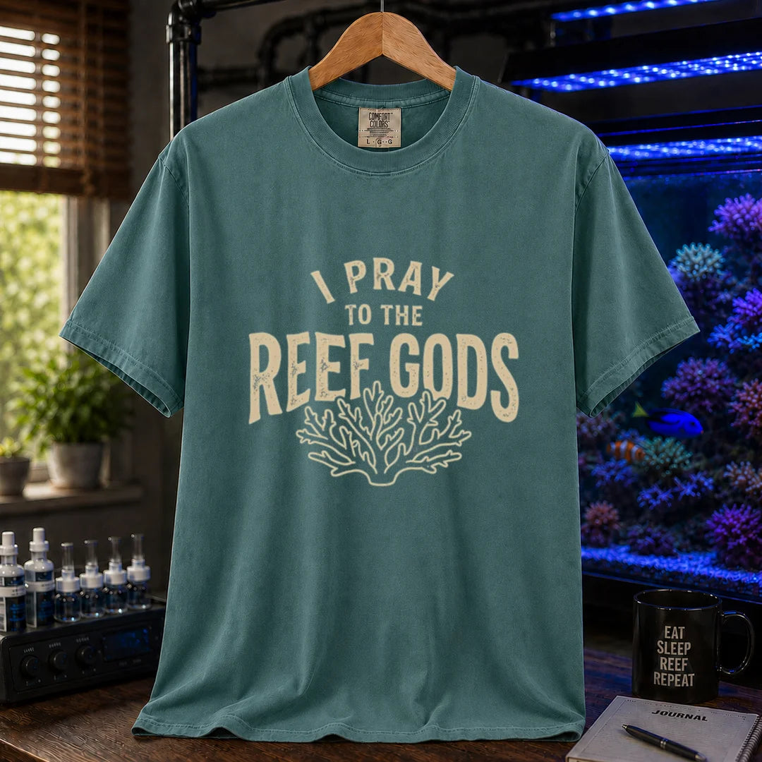 Saltwater aquarium graphic t-shirt featuring distressed cream typography reading I Pray To The Reef Gods with a coral line art graphic printed on a green tee displayed in a reef room setting