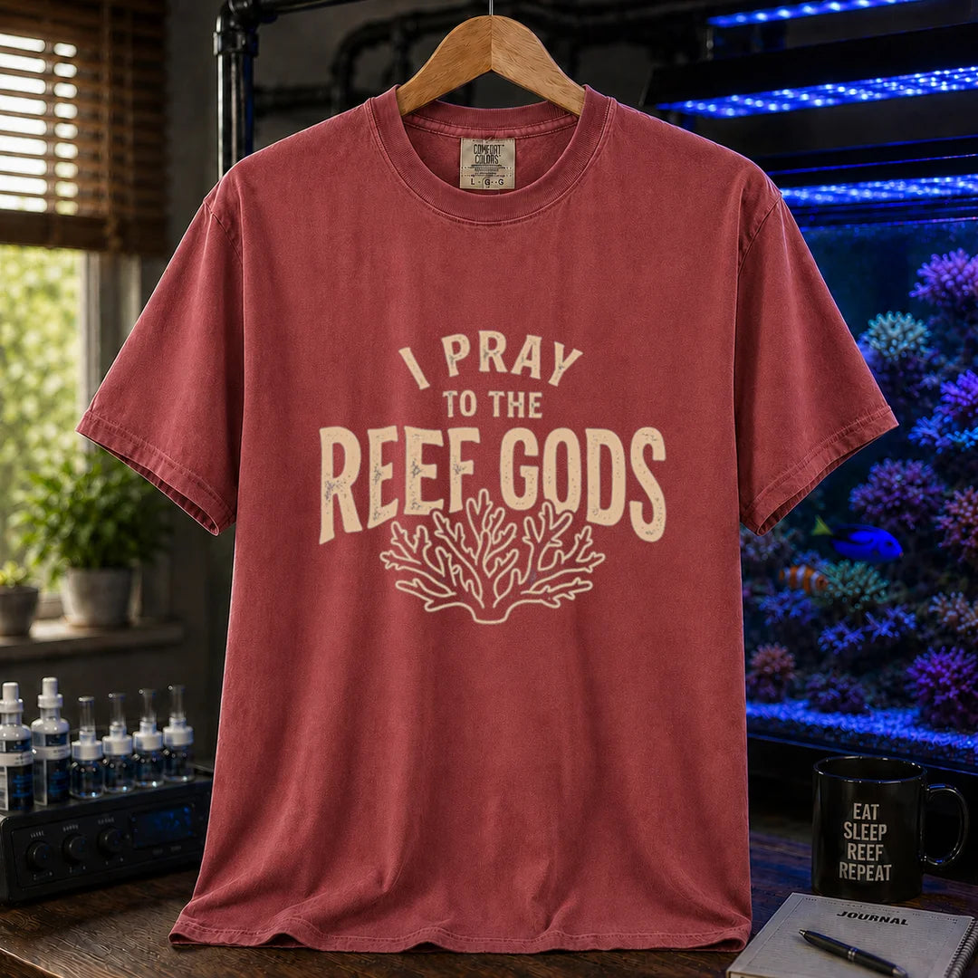 Saltwater aquarium graphic t-shirt featuring distressed cream typography reading I Pray To The Reef Gods with a coral line art graphic printed on a maroon, red tee displayed in a reef room setting