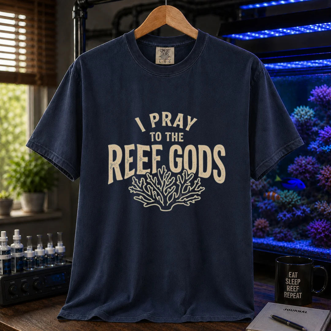 Saltwater aquarium graphic t-shirt featuring distressed cream typography reading I Pray To The Reef Gods with a coral line art graphic printed on a navy tee displayed in a reef room setting