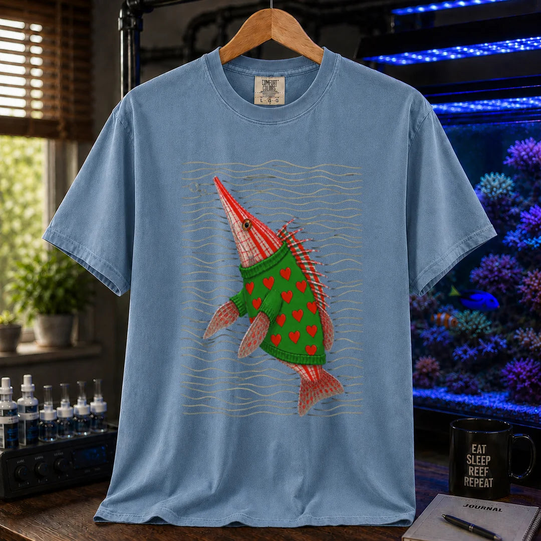 Saltwater aquarium graphic t-shirt featuring a whimsical illustration of a hawkfish wearing a green sweater with red hearts against wavy lines printed on a blue tee displayed in a reef room setting