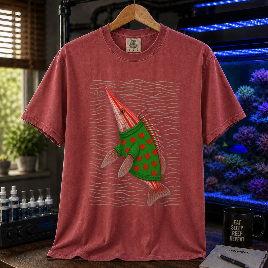 Saltwater aquarium graphic t-shirt featuring a whimsical illustration of a hawkfish wearing a green sweater with red hearts against wavy lines printed on a maroon, red tee displayed in a reef room setting