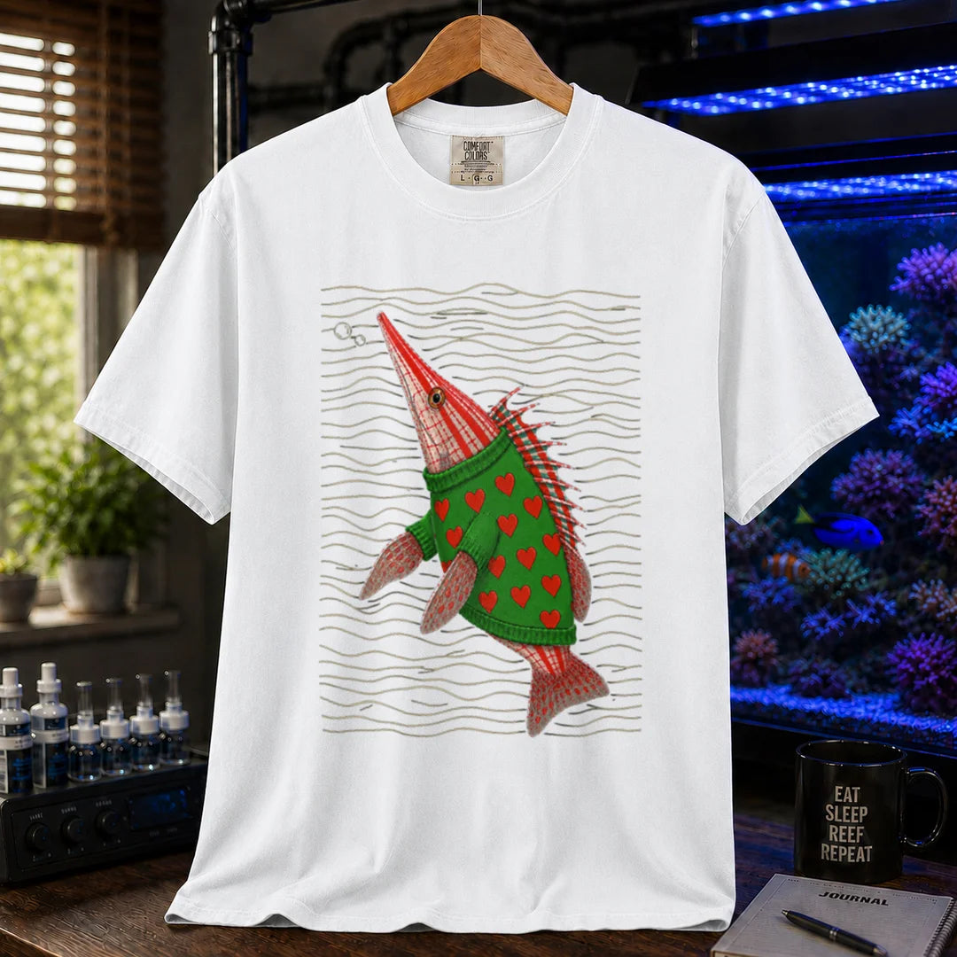 Saltwater aquarium graphic t-shirt featuring a whimsical illustration of a hawkfish wearing a green sweater with red hearts against wavy lines printed on a white tee displayed in a reef room setting
