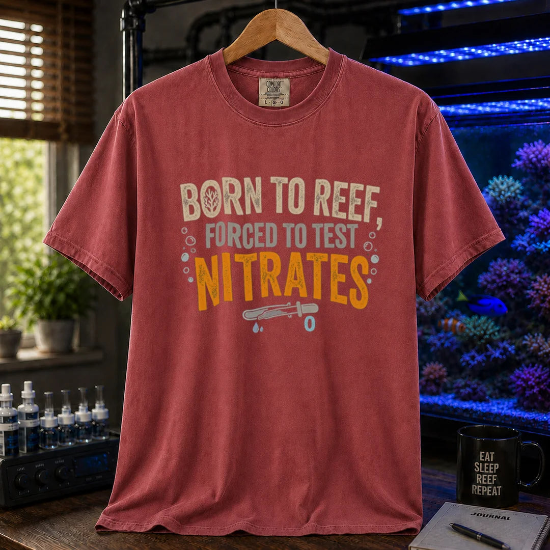 Saltwater aquarium graphic t-shirt featuring distressed cream, gray, and yellow-orange typography reading Born To Reef, Forced To Test Nitrates with a test vial icon printed on a maroon, red tee displayed in a reef room setting