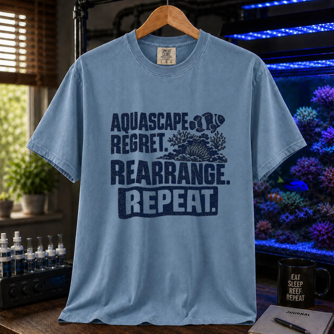 Saltwater aquarium graphic t-shirt featuring distressed dark blue typography reading Aquascape. Regret. Rearrange. Repeat. with a coral and clownfish illustration printed on a blue tee displayed in a reef room setting