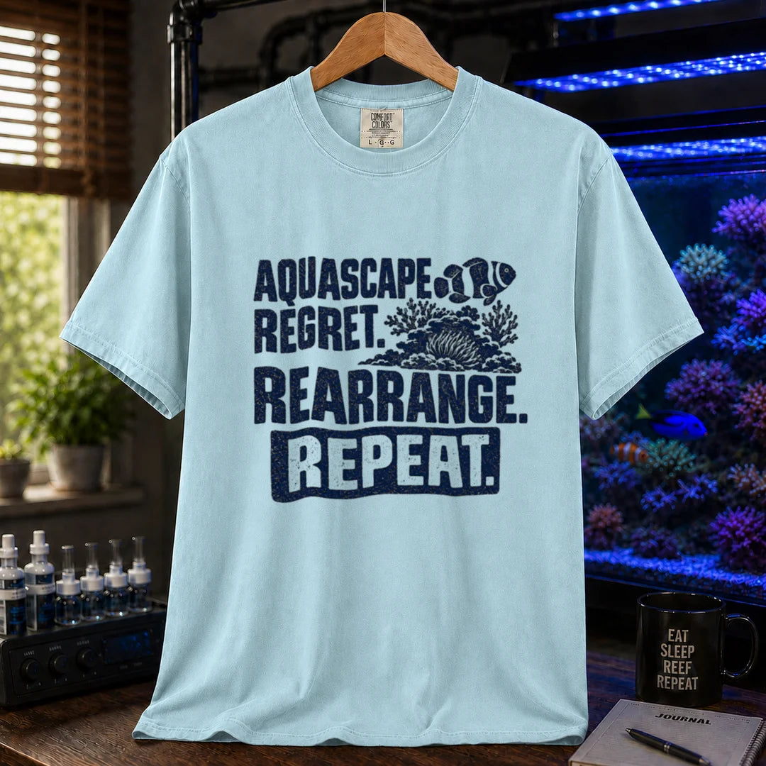 Saltwater aquarium graphic t-shirt featuring distressed dark blue typography reading Aquascape. Regret. Rearrange. Repeat. with a coral and clownfish illustration printed on a light blue tee displayed in a reef room setting