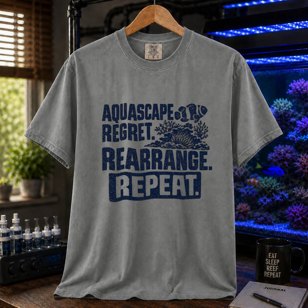 Saltwater aquarium graphic t-shirt featuring distressed dark blue typography reading Aquascape. Regret. Rearrange. Repeat. with a coral and clownfish illustration printed on a grey tee displayed in a reef room setting