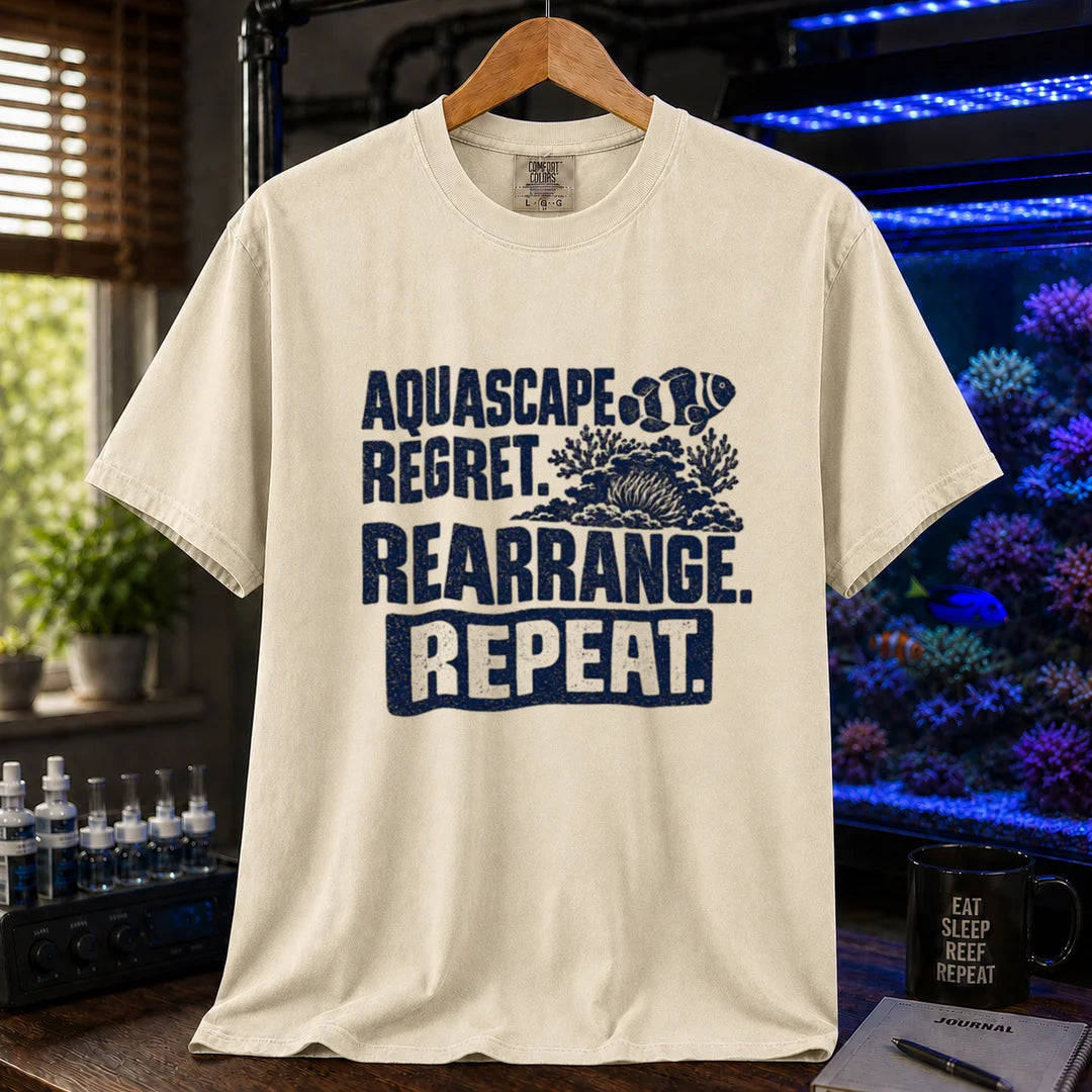 Saltwater aquarium graphic t-shirt featuring distressed dark blue typography reading Aquascape. Regret. Rearrange. Repeat. with a coral and clownfish illustration printed on a beige, cream, ivory tee displayed in a reef room setting