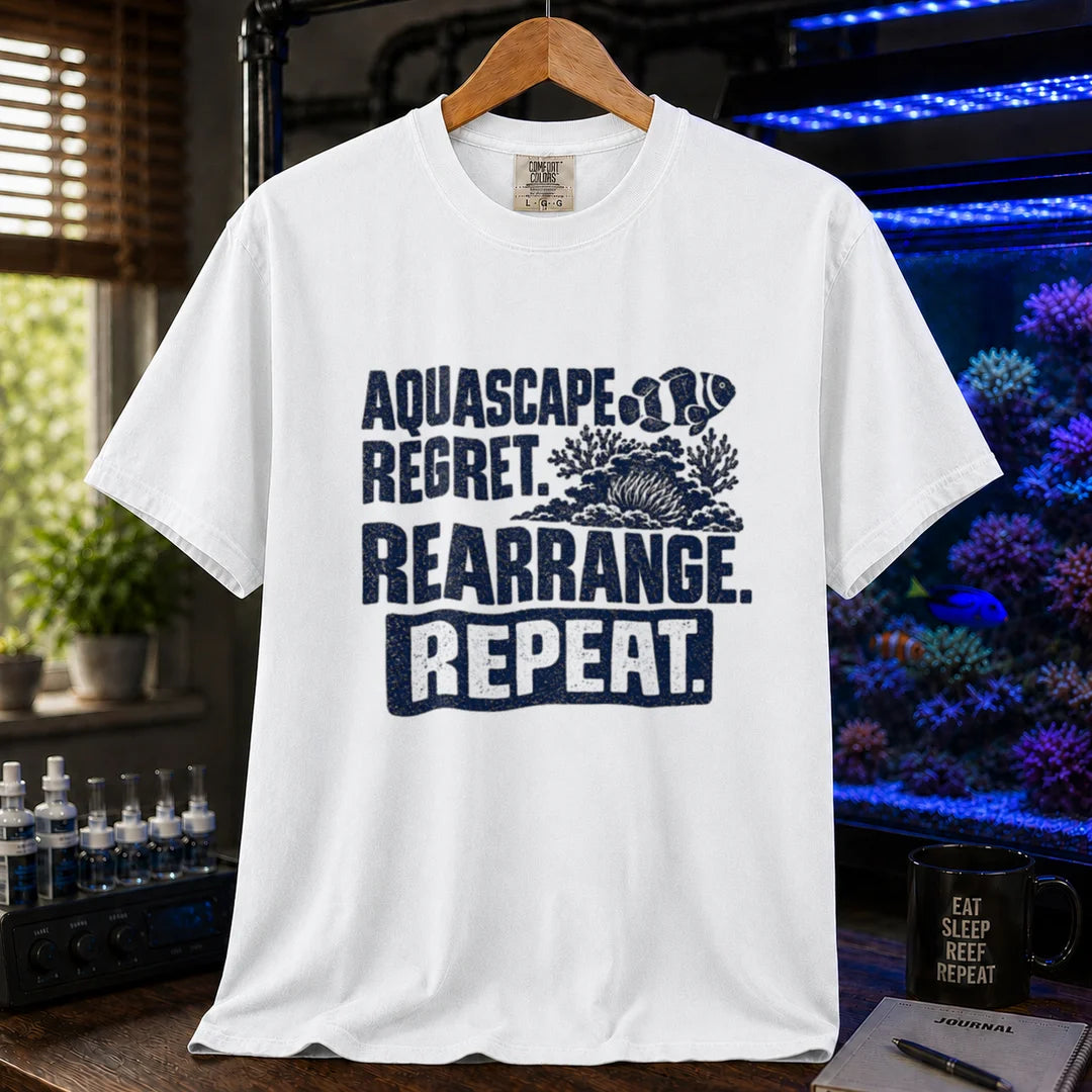 Saltwater aquarium graphic t-shirt featuring distressed dark blue typography reading Aquascape. Regret. Rearrange. Repeat. with a coral and clownfish illustration printed on a white tee displayed in a reef room setting